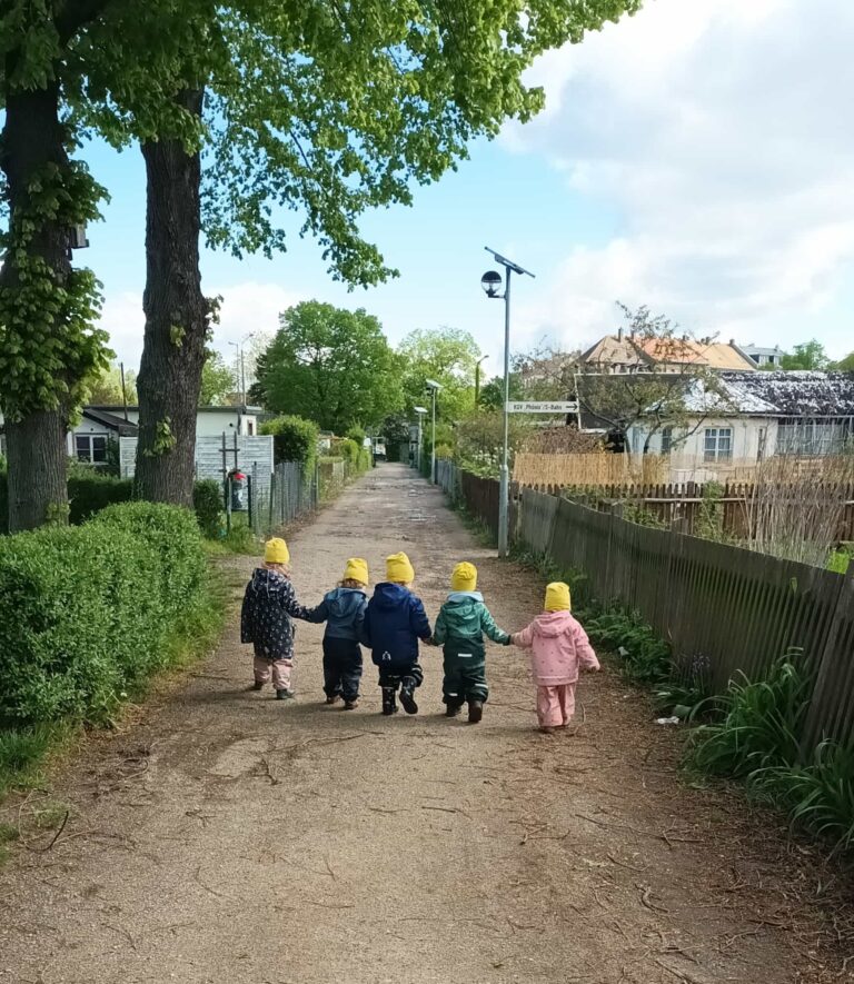 Kinder der Kindertagespflege nebeneinander an den Händen haltend laufen durch eine Gartenanlage